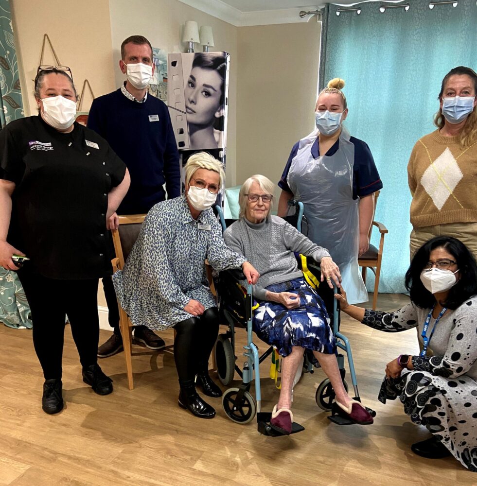 Image shows Dr Indira Kasibhatla crouching next to a female resident of Ashworth Grange care home with staff from the home standing around them. They're all wearing masks.