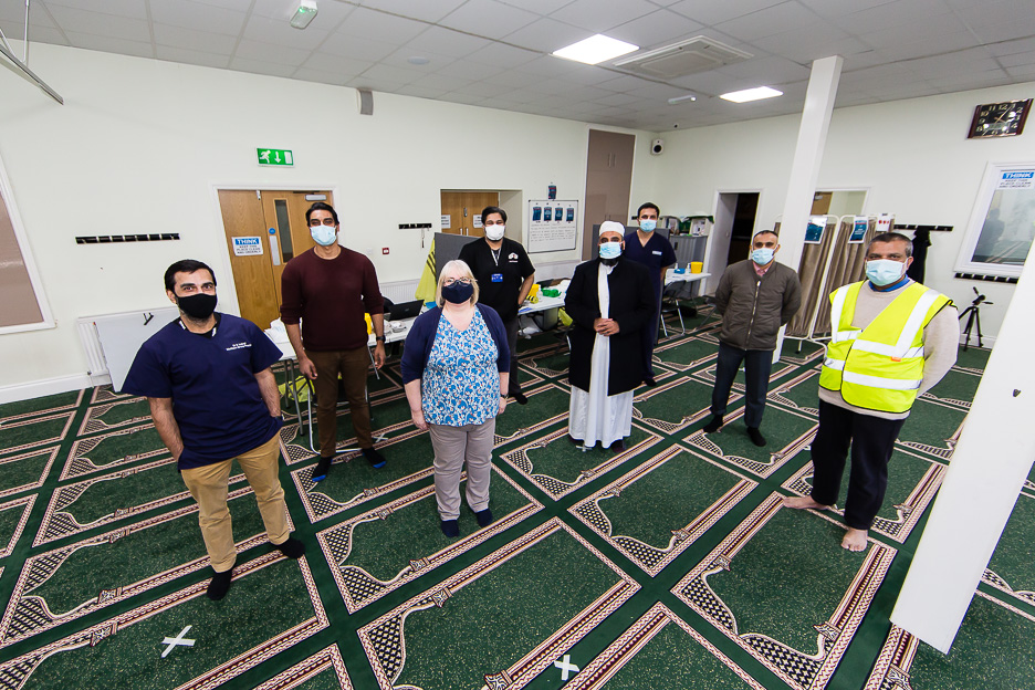 Eight people stood socially distanced. Three desks are behind them. They are all wearing face coverings. One man is wearing a high vis jacket.
