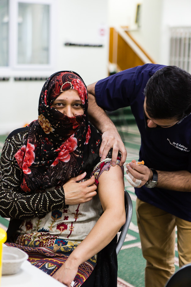 Woman wearing head scarf receives vaccine in left arm. She is sat down
