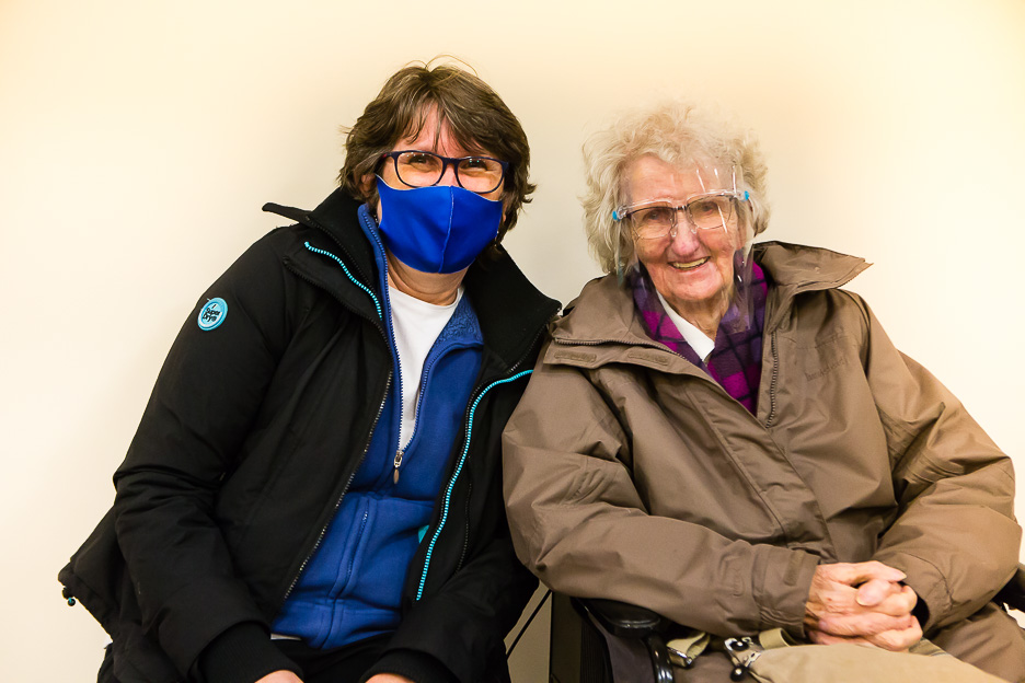 Two women sitting next to each other. The older one is wearing a visor. The younger one is wearing a mask.